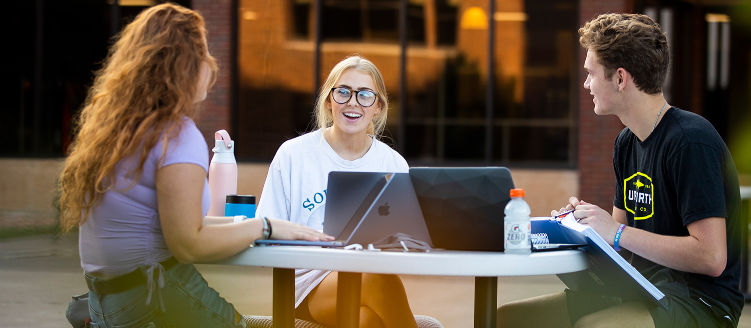 Three students studying together at a table outdoors while laughing.
