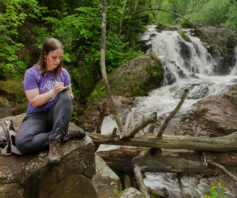 female student making notes while sitting on rock with stream and waterfall in background.