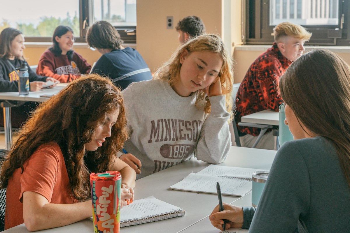 Three UMD students studying at a table in a bright, naturally lit classroom, with other students working at nearby tables in the background.