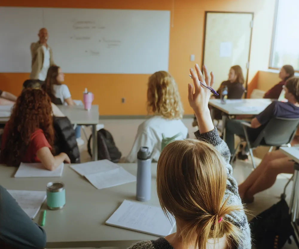 Students with back to camera in classroom raising hand.