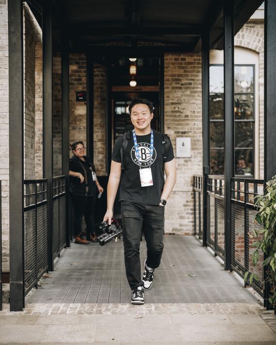 Tony Eaton walking along a covered patio outside of a building, carrying a camera