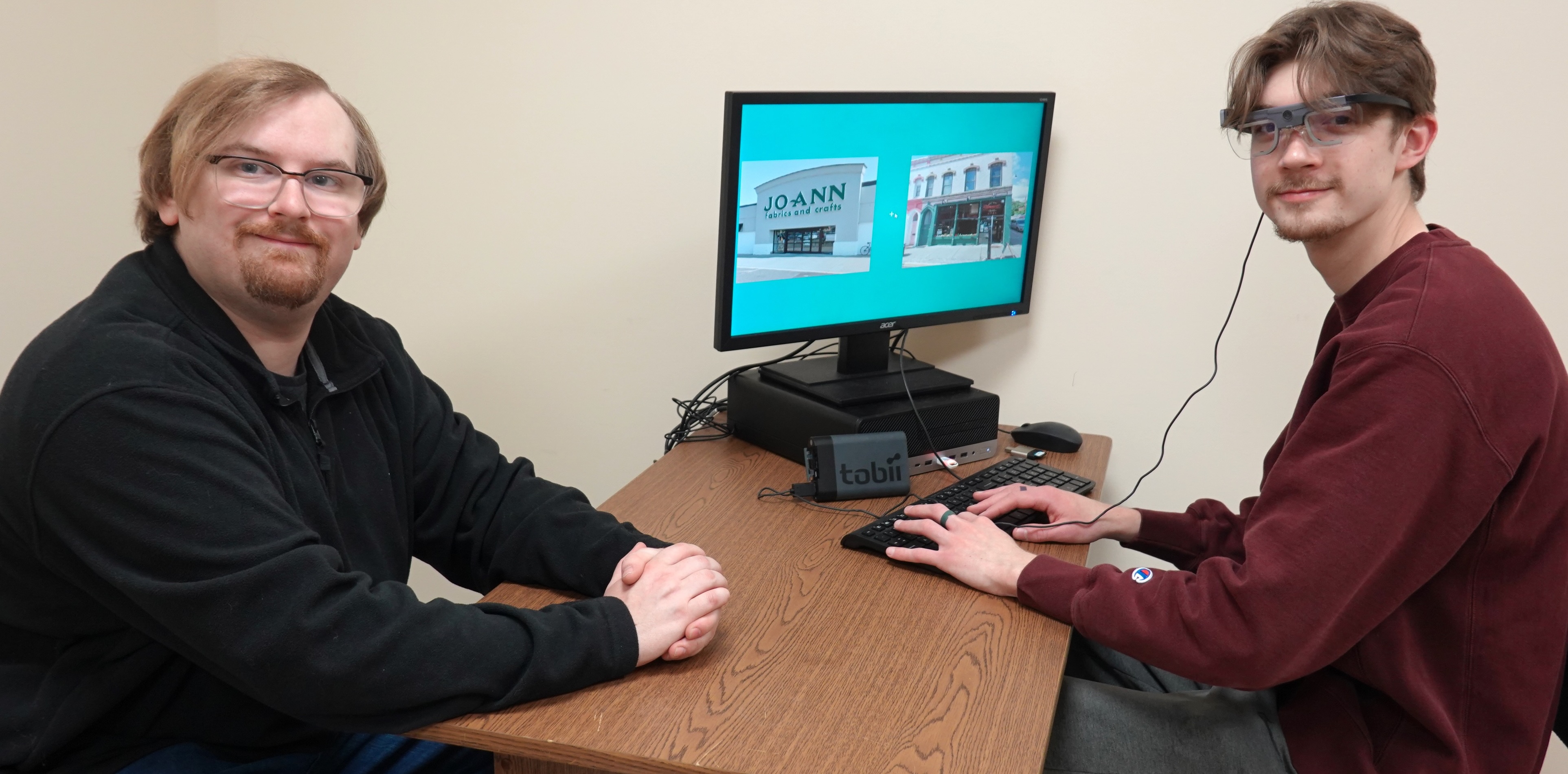 Two students sitting at a desk, one wearing eye tracking glasses with a computer screen in front of him