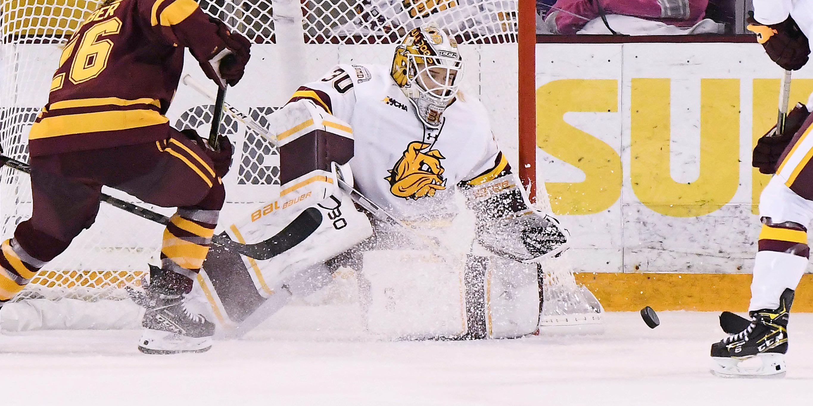A telephoto perspective showing a hockey goalie deflecting a puck in front of the net in AMSOIL Arena as two players skate in front of her.